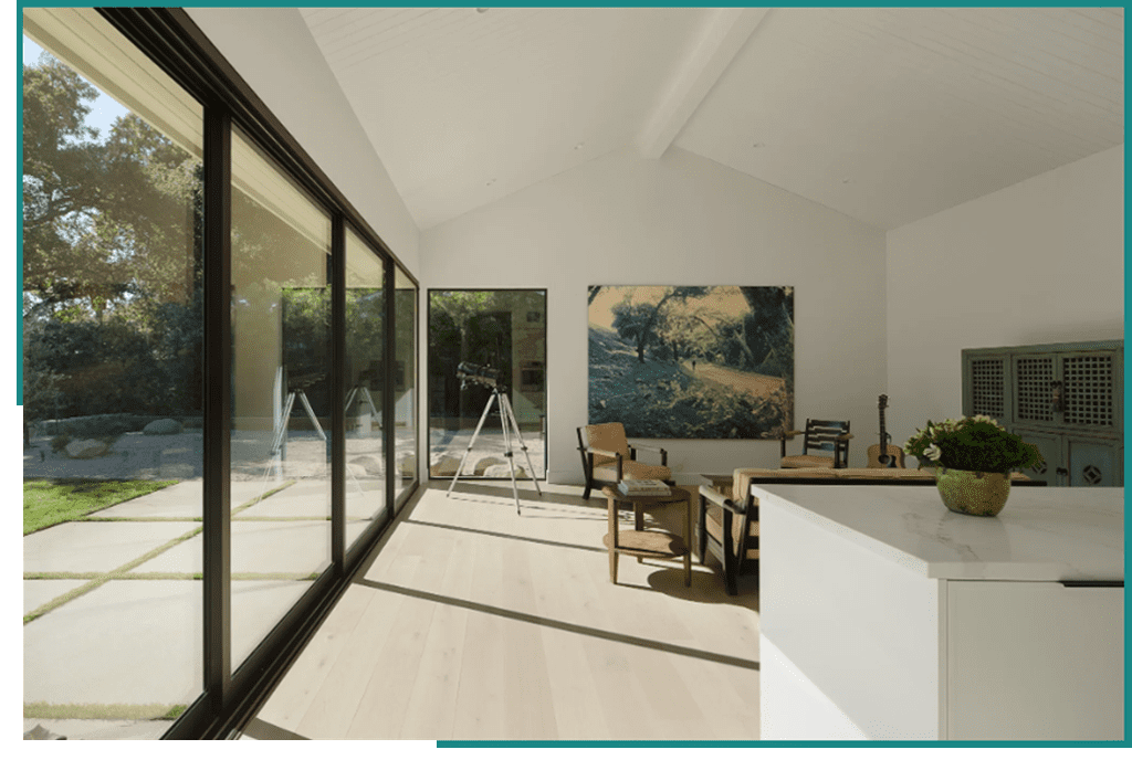 A spacious, modern room with a high white ceiling, large glass windows showcasing an outdoor view, wooden flooring, a simple chair with a footrest by an easel, artwork on the wall, and.