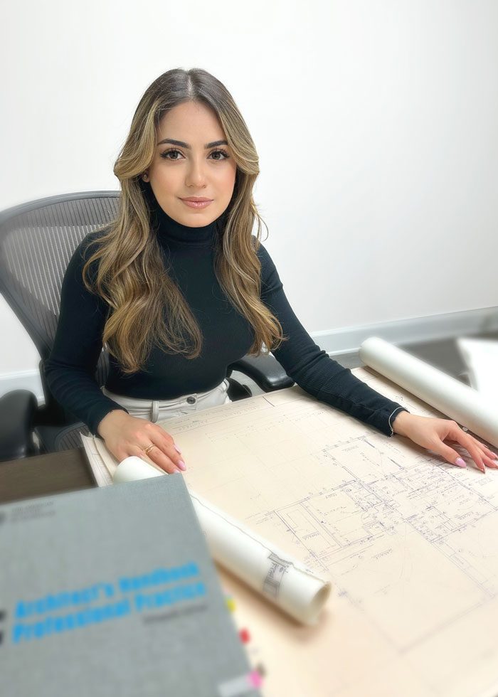 A woman in a black turtleneck sitting at a desk with blueprints and an engineering textbook, looking at the camera.