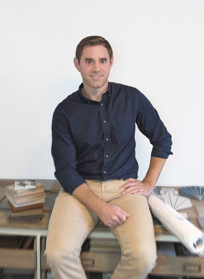 Man in a dark blue shirt and beige pants sitting on a desk, smiling at the camera, with books and plans beside him.